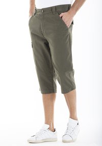 Shorts cargo vert olive en tissu léger, arrivant aux genoux avec des poches latérales, portés avec des baskets blanches. Dotés d'une fermeture à boutons.