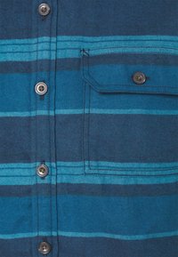 Camicia con bottoni caratterizzata da righe orizzontali blu scuro e turchese, con taschino e bottoni color argento. Texture del tessuto visibile.