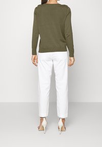 Maglione a maglia verde oliva con scollatura a barca, polsini e orlo a costine, abbinato a pantaloni bianchi a gamba affusolata e scarpe con tacco color crema.