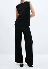 Žena v bezrukávovém černém žebrovaném topu a širokých kalhotách, doplněná černými špičatými vysokými podpatky s dekorativními přezkami.