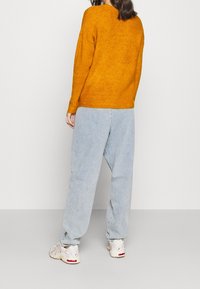 Senapsgul stickad tröja med rundad halsringning, kombinerad med ljusblå vida jeans. Vita sneakers med röda detaljer kompletterar outfiten.