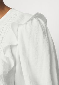Blusa branca com mangas franzidas, apresentando detalhes em renda ao longo da gola. Feita de um tecido suave e leve com uma superfície ligeiramente texturizada.