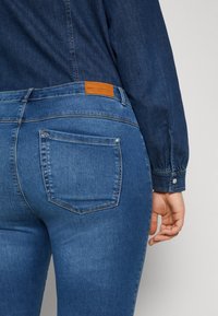 Detailní záběr osoby, která má na sobě modré džínové kalhoty a tmavou džínovou košili, se zaměřením na zadní kapsu a koženou značkovou etiketu.