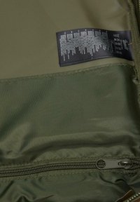 Oliņzaļš auduma soma ar gludu virsmu, aprīkota ar rāvējslēdzi un aprūpes etiķeti, uz kuras ir iespiestas lietošanas instrukcijas.