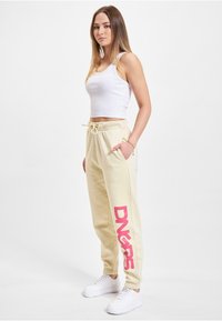 Helle gelbe Sweatpants mit einem rosa Logo, auf dem "DNRS" steht, mit elastischem Bund, Kordelzug und seitlichen Taschen, kombiniert mit weißen Sneakers.