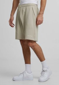 Lichtbeige, getextureerde shorts met een elastische tailleband, voorzien van zijzakken en een klein wit logo op de zoom, gecombineerd met witte sneakers.