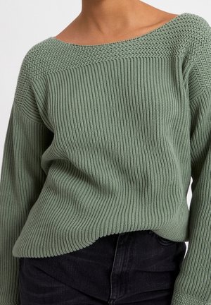 Světlé zelený pletený svetr s žebrovanými a texturovanými vzory, nošený s černými kalhotami, zkrácený v pase, viditelné jsou ramena.