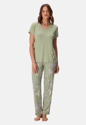 Vrouw staat op blote voeten, draagt een lichtgroen shirt met korte mouwen en bijpassende losse broek met bloemenpatroon op een effen witte achtergrond.