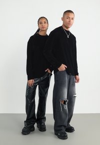 Două modele pozează. Primul poartă un top lung cu mânecă din catifea neagră și pantaloni din piele lucioasă negri. Al doilea poartă un top lung cu mânecă negru și blugi gri uzat.