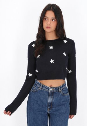 Ung kvinde iført en sort cropped sweater med hvide blomstermønstre og højtaljede blå jeans, kigger til siden mod en hvid baggrund.