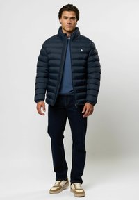 Mees, kes kannab tumesinist puffer-jopet, sinist kampsunit, tumedaid teksapükse ja beež-valgeid vabaajajalatseid, seisab ühetaustalise heleda tausta ees.