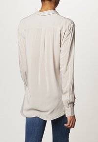 Vaaleanbeige pitkähiha paita, jossa on kaulus, tehty sileästä kankaasta, takana laskos ja napilliset kalvosimet. Yhdistetty tummiin farkkuihin.