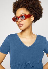 Top de algodão azul com decote em V, mangas curtas e cintura com cordão. Óculos de sol oversized em padrão tartaruga vermelho com lentes azuladas.
