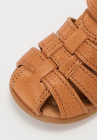 Sandale en cuir tan avec un design tissé, silhouette ouverte sur les orteils et surpiqûres contrastées. Semelle en caoutchouc plate, sans embellissements.