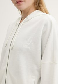 Sweat à capuche blanc avec fermeture éclair, arborant un logo de cœur brodé. Le matériau est doux au toucher et possède des poches latérales.