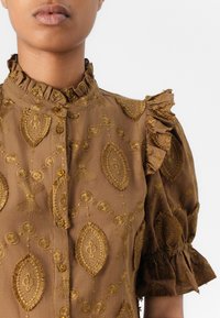 Blusa marrone ricamata con collo a balze e maniche a palloncino. Presenta una decorazione con cuciture dorate in motivi intricati e chiusura con bottoni.