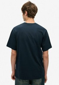 Man in een effen donkerblauwe kortemouwshirt en spijkerbroek, staande met zijn rug naar de camera tegen een witte achtergrond.