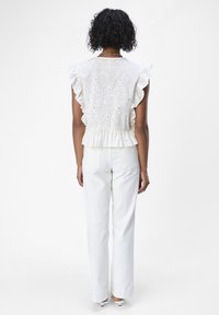 Hvid, klokkeformet top med broderet detaljer og peplum talje, parret med hvide lige bukser. Modellen vises fra bagsiden.