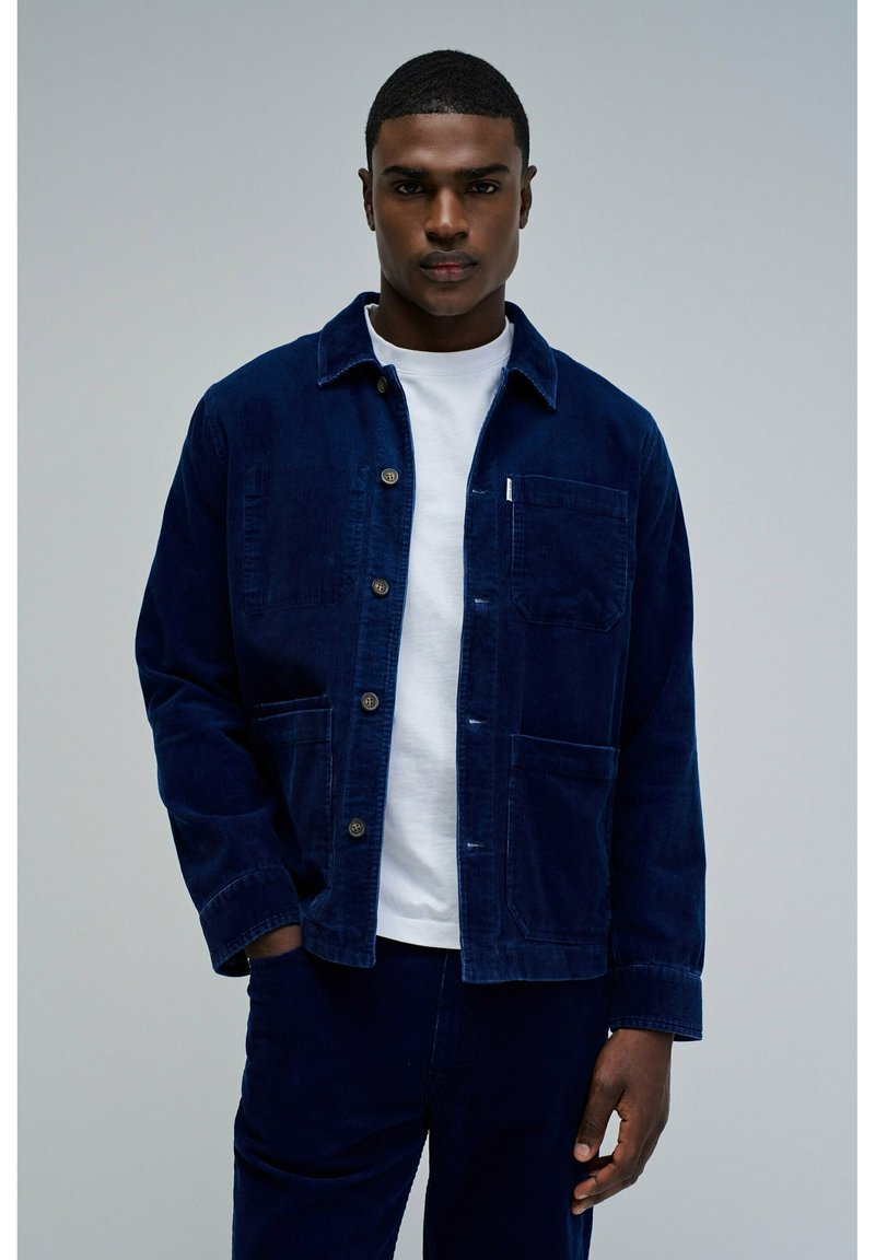 Veste en velours côtelé bleu marine avec devant à boutons, deux poches avant et un col plié, portée sur un t-shirt blanc. Texture lisse, coupe décontractée.