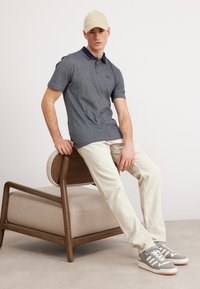 Pelēka polo krekla ar tumši zilu apkakli, beiges cepures, krēmkrāsas bikses un pelēkiem sporta apaviem, kas sēž uz koka krēsla ar mīkstu sēdekli.