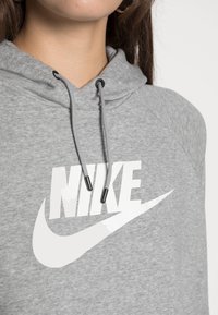 Närbild på en person som bär en grå hoodie med gråa dragsnören och en stor vit Nike-logotyp på bröstet.