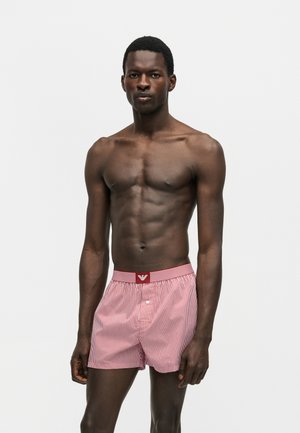 Atletyczny ciemnoskóry mężczyzna stoi prosto, nosząc czerwono-białe w paski bokserki z guzikiem i logo na pasie, na jednolitym tle.