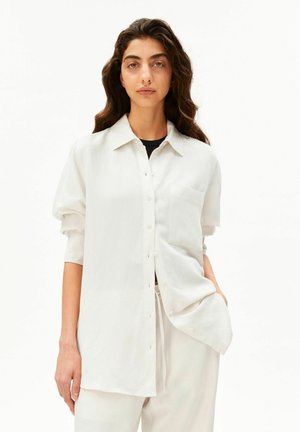 Frau mit langen, dunklen, welligen Haaren, die ein lockeres weißes Hemd mit Knöpfen und passende weiße Hosen trägt, stehend vor einem schlichten weißen Hintergrund.