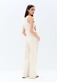 Tuta beige con pantaloni larghi in un tessuto leggero, caratterizzata da un design senza maniche, tasche laterali e uno scollo a V sul retro. Stile pulito e minimalista.