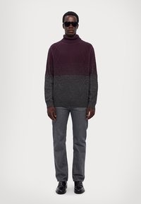 Pull col roulé bordeaux avec un motif géométrique texturé, dégradé vers le gris. Associé avec un jean gris et des chaussures noires, accessoirisé avec des lunettes de soleil.