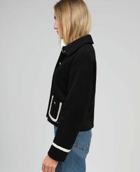 Femme aux cheveux blonds portant une veste noire à boutons avec des bordures blanches sur les poches et les manches, associée à un jean bleu, faisant face à gauche.