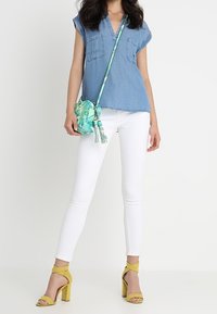 Top azzurro di denim a maniche corte con due tasche sul petto, jeans skinny bianchi e décolleté con cinturino giallo; borsa a tracolla con motivo verde.