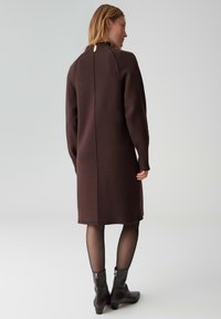 Bruine, gebreide jurk met lange mouwen, tot halverwege de kuit, met een split aan de achterkant en een hoge hals. Draagt zwarte enkelhoge laarzen.