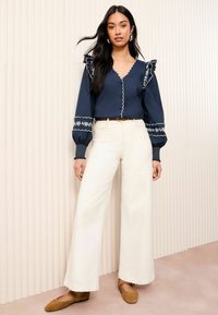 Navy blouse with scalloped neckline and embroidered accents, paired with wide-leg white trousers and brown flats. Textured background.