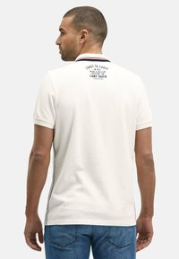 Man met kort haar in een witte poloshirt met tekst op de bovenrug en blauwe spijkerbroek, staand tegen een effen achtergrond.