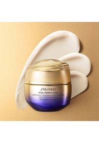Shiseido Vital Perfection crème in een ronde pot met een gouden deksel. Een gladde witte crèmemonster ernaast op een gestructureerde gouden achtergrond.