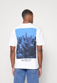Witte t-shirt met een blauw en zwart stadsbeeld van New York City op de achterkant, met de tekst "NEW YORK CITY QUEENS BOROUGH."