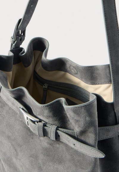 Sac fourre-tout en suède gris avec un bord supérieur froncé, une bandoulière réglable et un intérieur crème avec une poche zippée. Accents en métal argenté.