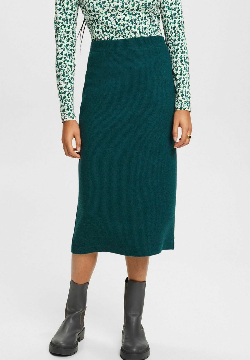 Femme portant une jupe tricotée bleu sarcelle jusqu'aux genoux, un haut à manches longues à motifs verts et blancs, et des bottines noires, debout sur un fond uni.