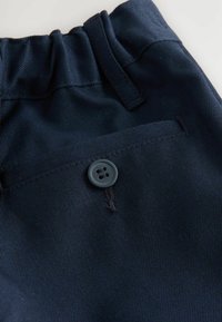 Navyblauem Stoff mit strukturierter Oberfläche, ausgestattet mit einem elastischen Bund und einem einzelnen grauen Knopf an einer flachen Tasche. Minimale Nähdetails.