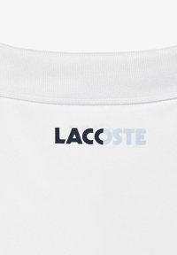 Col un tissu blanc avec le logo "LACOSTE" imprimé en noir et en bleu clair sous l'encolure.