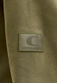 Tessuto verde oliva con superficie testurizzata, caratterizzato da una toppa logo rettangolare in rilievo dello stesso colore. Il testo recita "JACK & JONES."