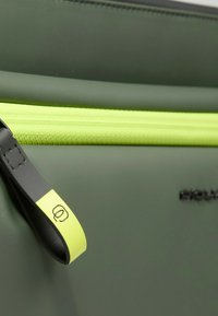 Bolsa verde con textura suave, que presenta una cremallera amarilla brillante y una lengüeta de tirón, además de un detalle de marca. Colores contrastantes prominentes.