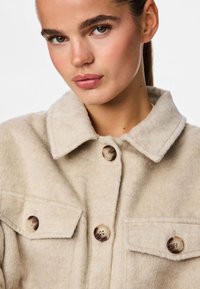 Beige jacka i ullblandning med texturerad yta, spetsig krage, två fickor med lock och stora runda knappar i kontrasterande färger.