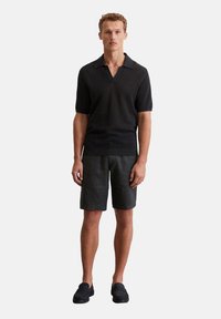 Schwarzes, kurzärmliges Poloshirt mit V-Ausschnitt, kombiniert mit grauen Shorts. Das Model trägt dunkle Slipper und steht vor einem schlichten Hintergrund.