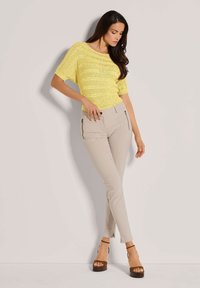 Gelbes, locker verstricktes Oberteil kombiniert mit schmal geschnittenen beigen Hosen mit Reißverschlusstaschen, ergänzt durch braune Absatzsandalen.