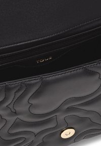 Borsa a mano in pelle nera con motivo ondulato trapuntato, hardware dorato, chiusura con zip e nome del brand "TOUS" impresso all'interno.