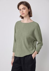 Top verde chiaro, testurizzato, con scollatura ampia e maniche a tre quarti; abbinato a pantaloni neri con tasche laterali.