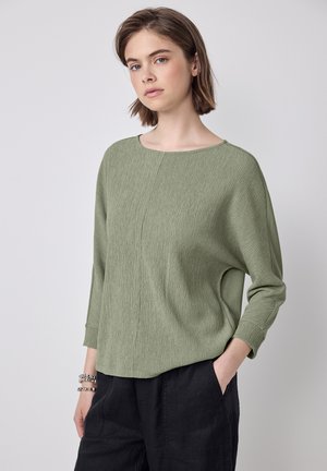 Top verde chiaro, testurizzato, con scollatura ampia e maniche a tre quarti; abbinato a pantaloni neri con tasche laterali.