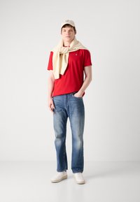 T-shirt rouge, jeans bleu clair, baskets blanches et un pull couleur crème drapé sur les épaules. Casquette beige avec un détail logo.