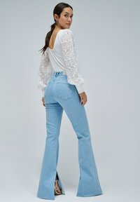 Top blanco con mangas de encaje abullonadas, combinado con jeans acampanados de color azul claro que tienen cortes en el dobladillo. Textura suave, cintura ajustada.